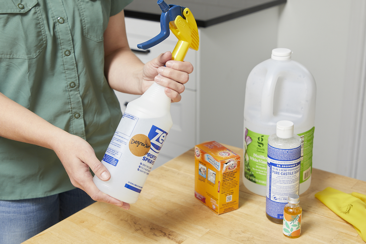 Une femme tient un vaporisateur étiqueté « dégraissant » avec des contenants de vinaigre et de bicarbonate de soude à proximité.