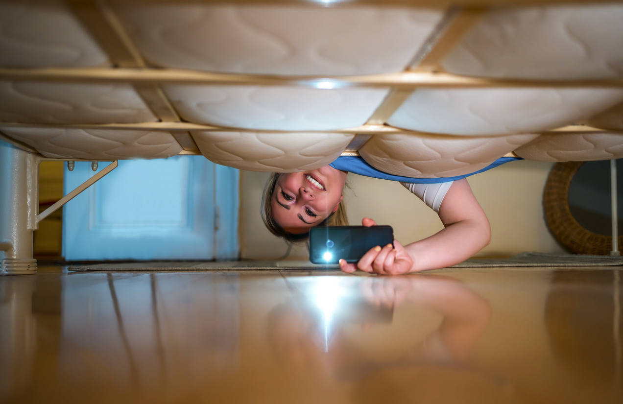 Jeune femme souriante regardant sous le lit, utilisant une lampe de poche à la recherche