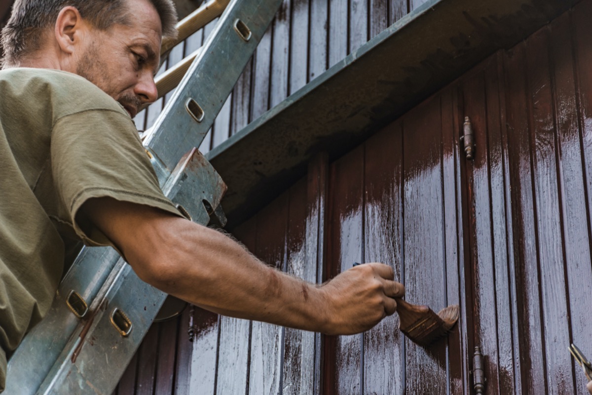 Man standing on a ladder leaning against a house, painting the house brown.