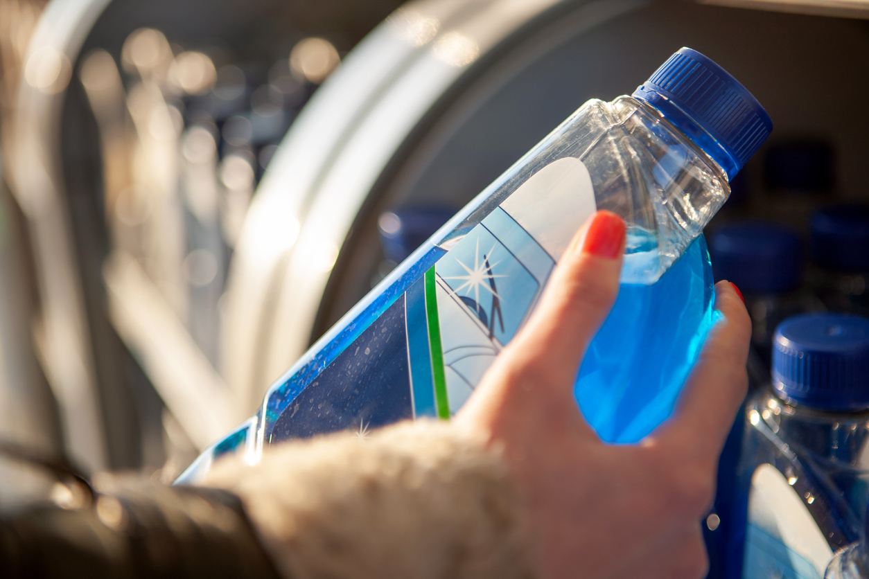 woman's hand with red nail holding a bottle of blue windshield wiper fluid in front of open car engine