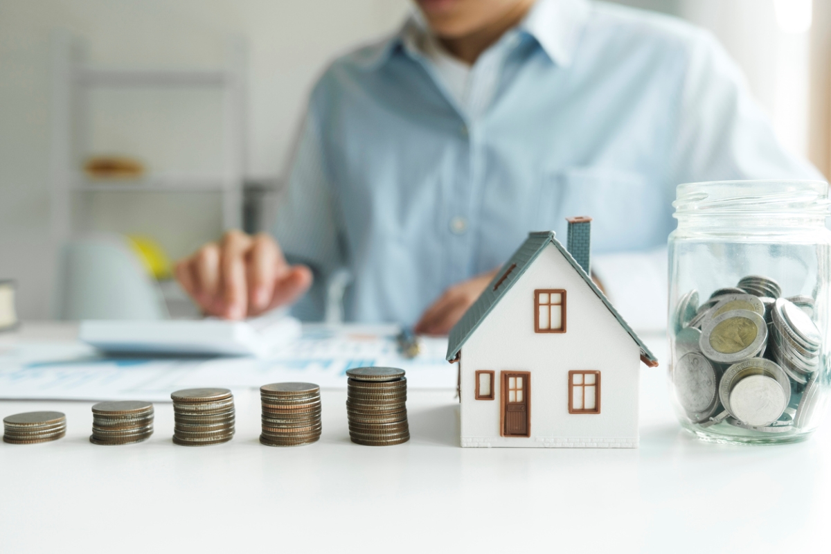 Stacked coins with model home and jar of coins on table.