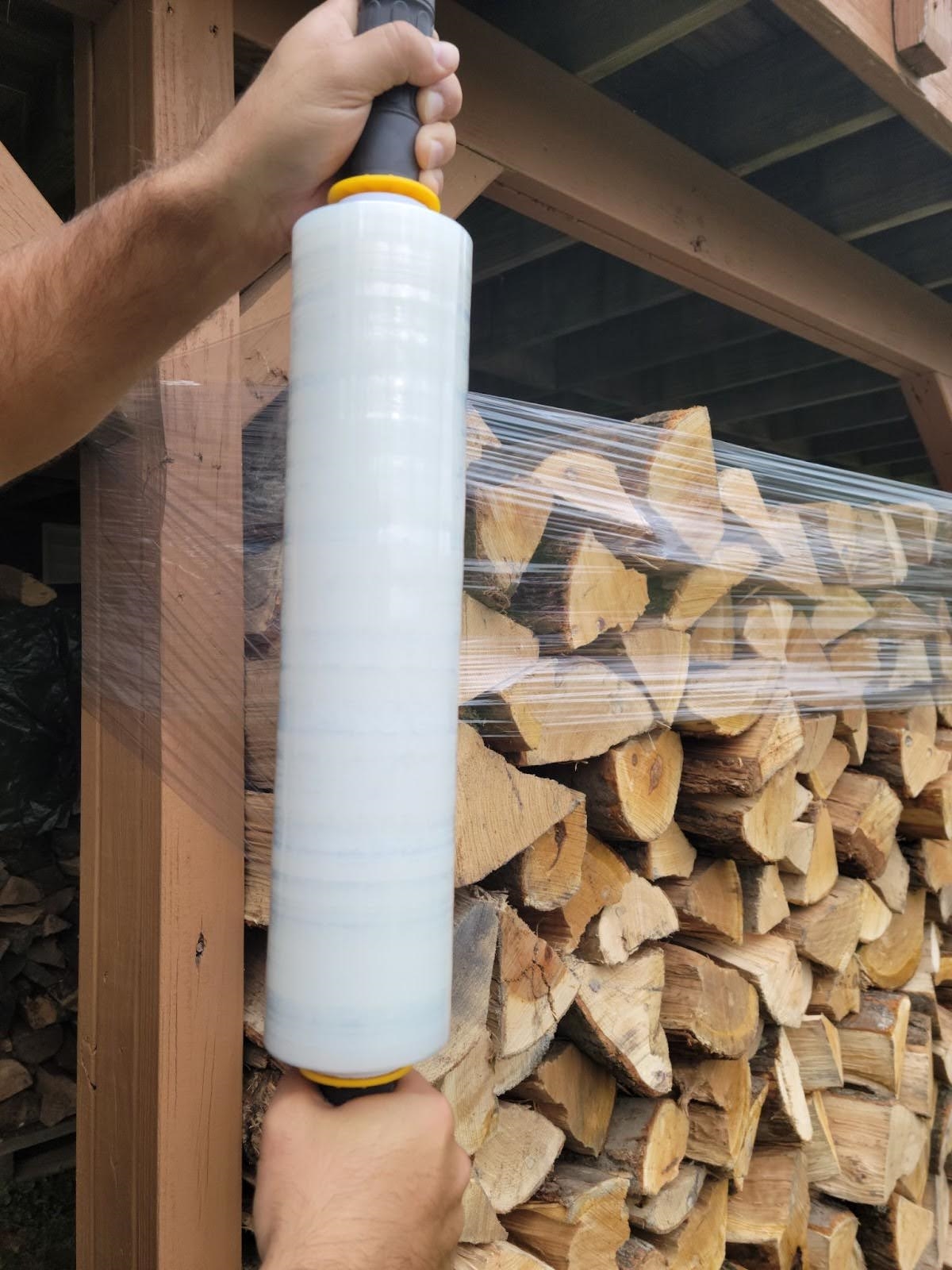 Person wrapping stack of firewood with large roll of plastic wrap.