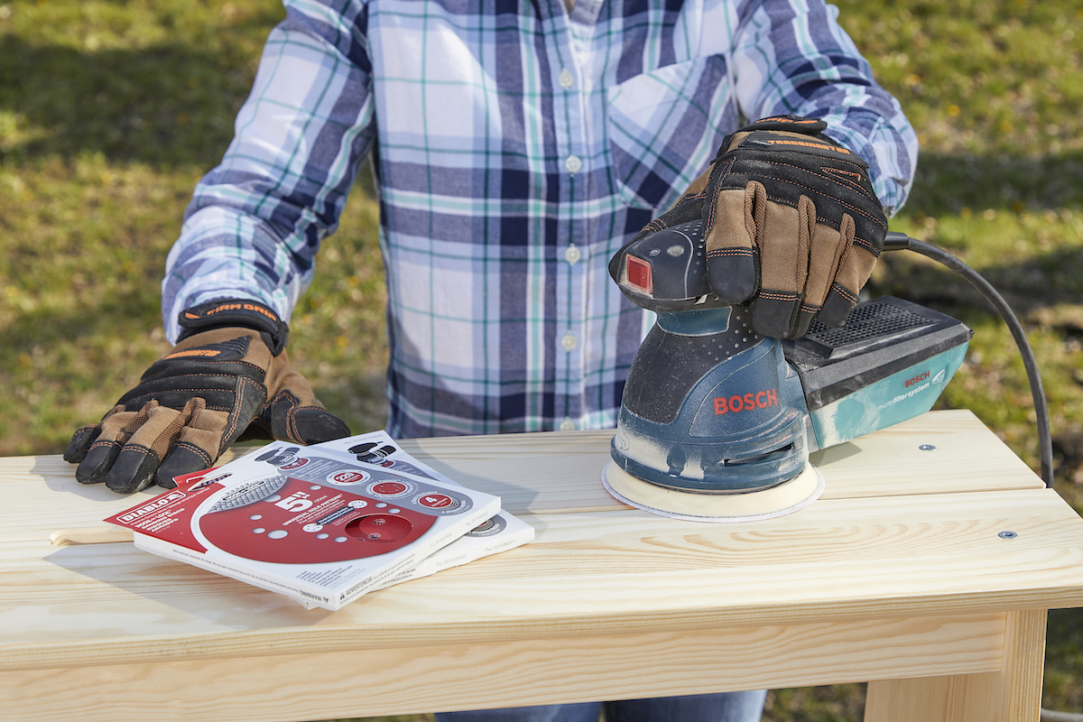 Woman has palm sander in one hand, with stacks of fine-grit sanding discs by the other hand.