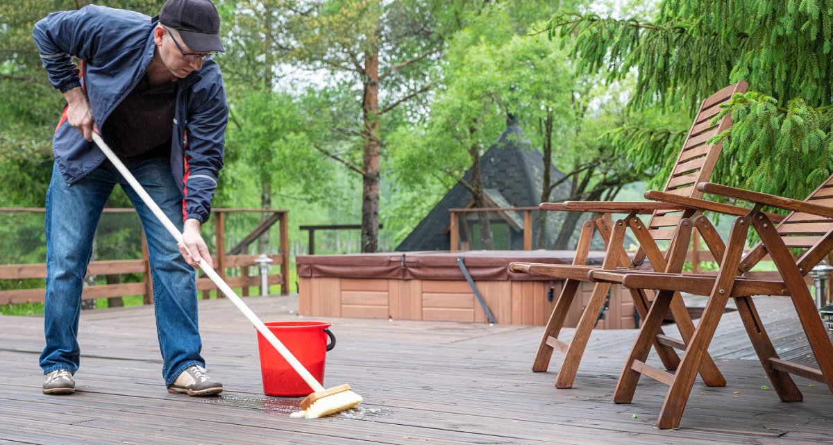 Une personne d'âge mûr est penchée en avant et frotte la surface d'une grande terrasse en bois avec une brosse. Un seau rouge contenant du liquide est posé juste à côté de lui.