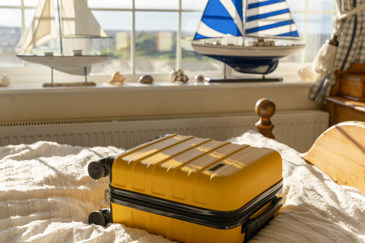 A yellow suitcase on a bed next to a large window with model sailboats on the windowsill.