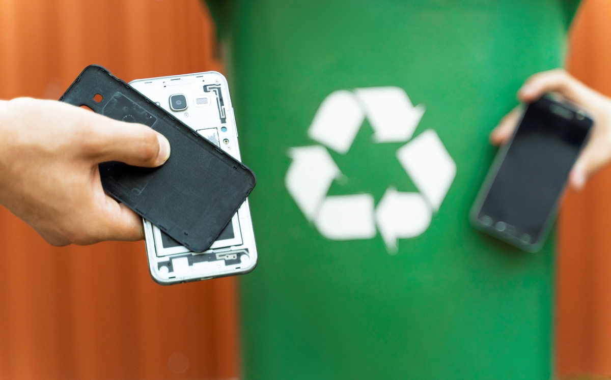 Hands holding old cellphones with green recycle bin in background