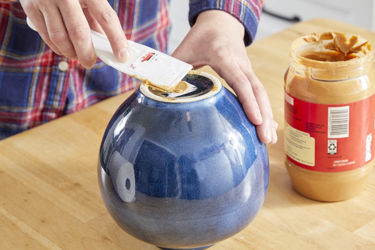 Woman spreads peanut butter on bottom of a blue vase to remove price sticker.