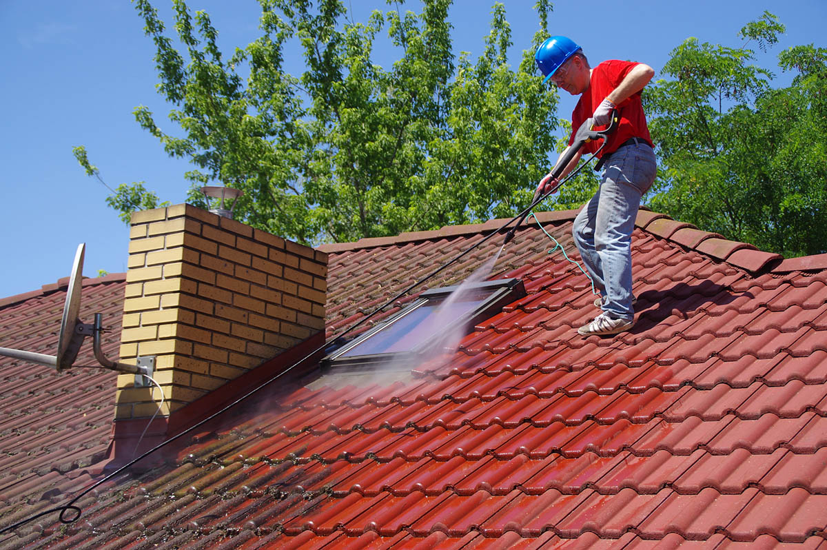 A worker wearing a hard hat cleans a roof using a power washer.