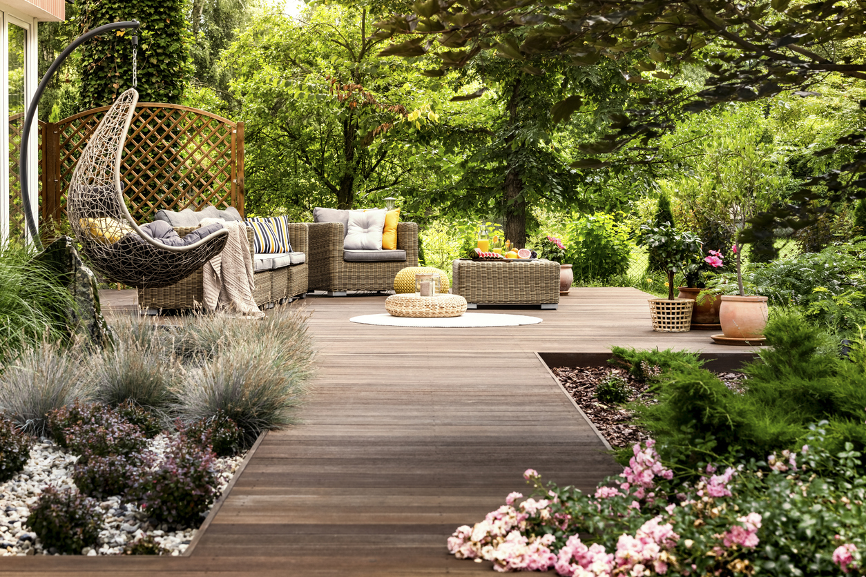 Une terrasse en bois entourée de verdure, avec des meubles de patio en osier et des graminées ornementales.