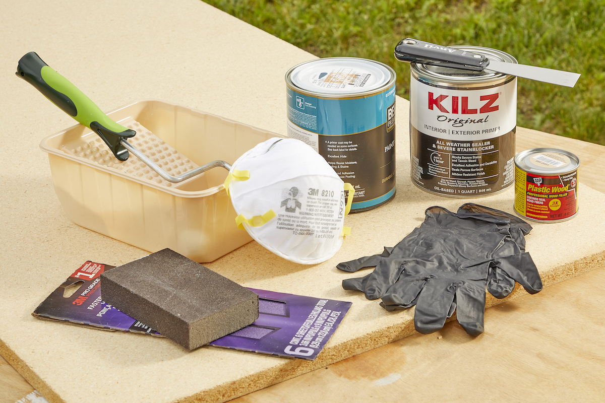 Materials needed to paint particle board including paint, roller, primer, and gloves, on an outdoor table.