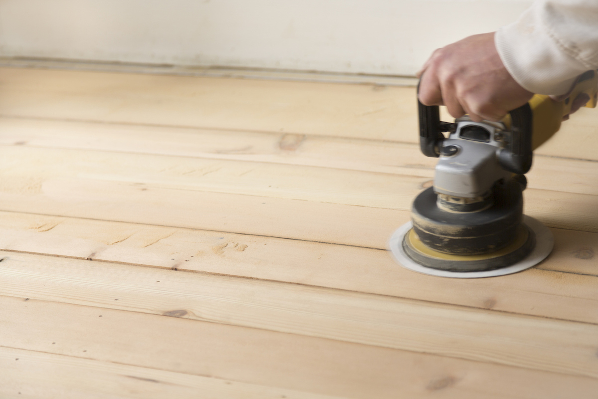 Man is using a random orbital sander to sand light-colored hardwood flooring.