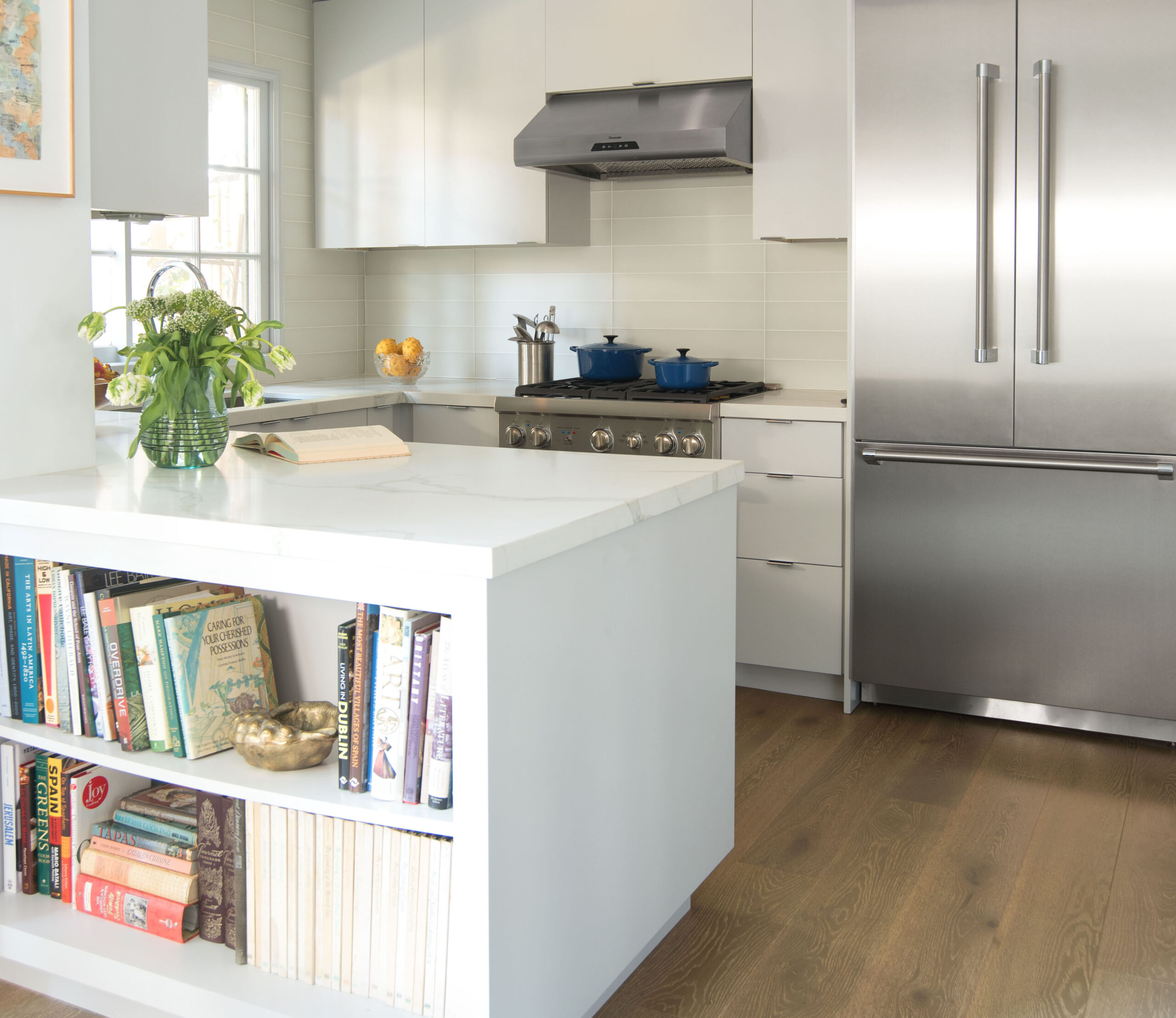 A wide view of a kitchen with a range hood over the stove and a large open shelving unit in the foreground filled with books.