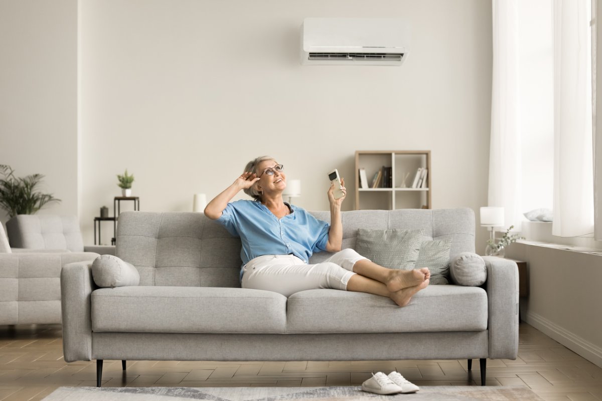 A woman sits on a grey couch holding a remote that controls the AC mounted on the white wall behind her. 