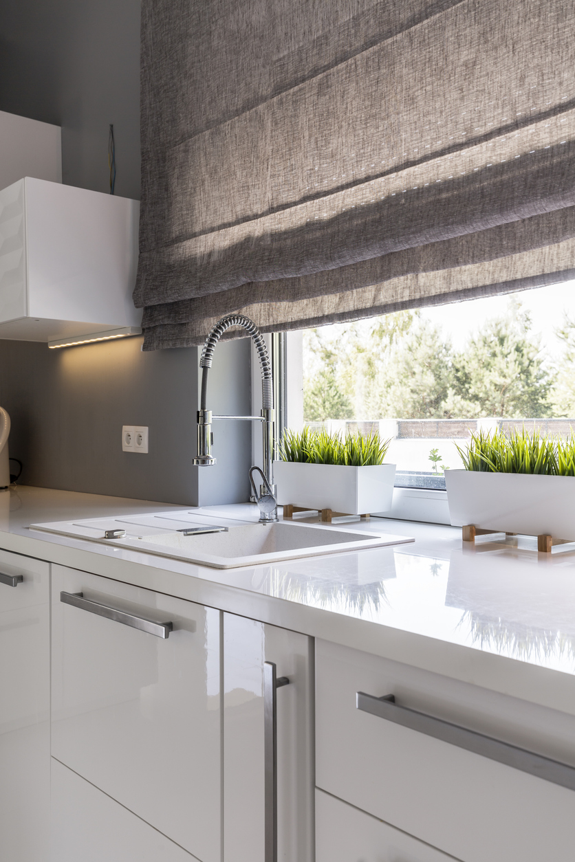 A small kitchen with large window covered by a gray roman shade.