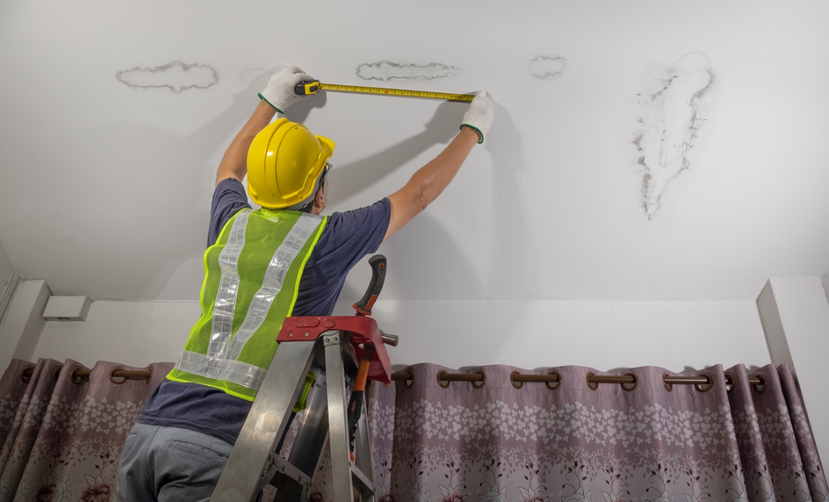 Professional on ladder using measuring tape on ceiling water stains.