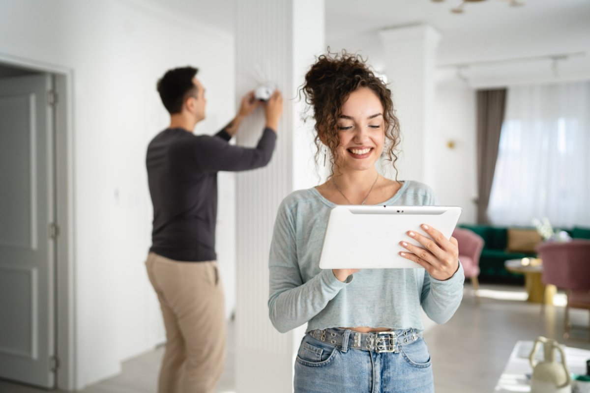 A woman smiles while using a smart tablet; a man behind her installs a home security system. 