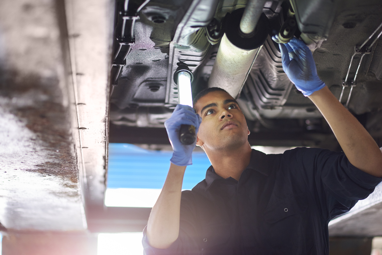 A young mechanic is working under a car in a garage repair shop. He is wearing blue overalls. He is looking up with an inspection lamp to check the possible damage that has been sustained to the exhaust.