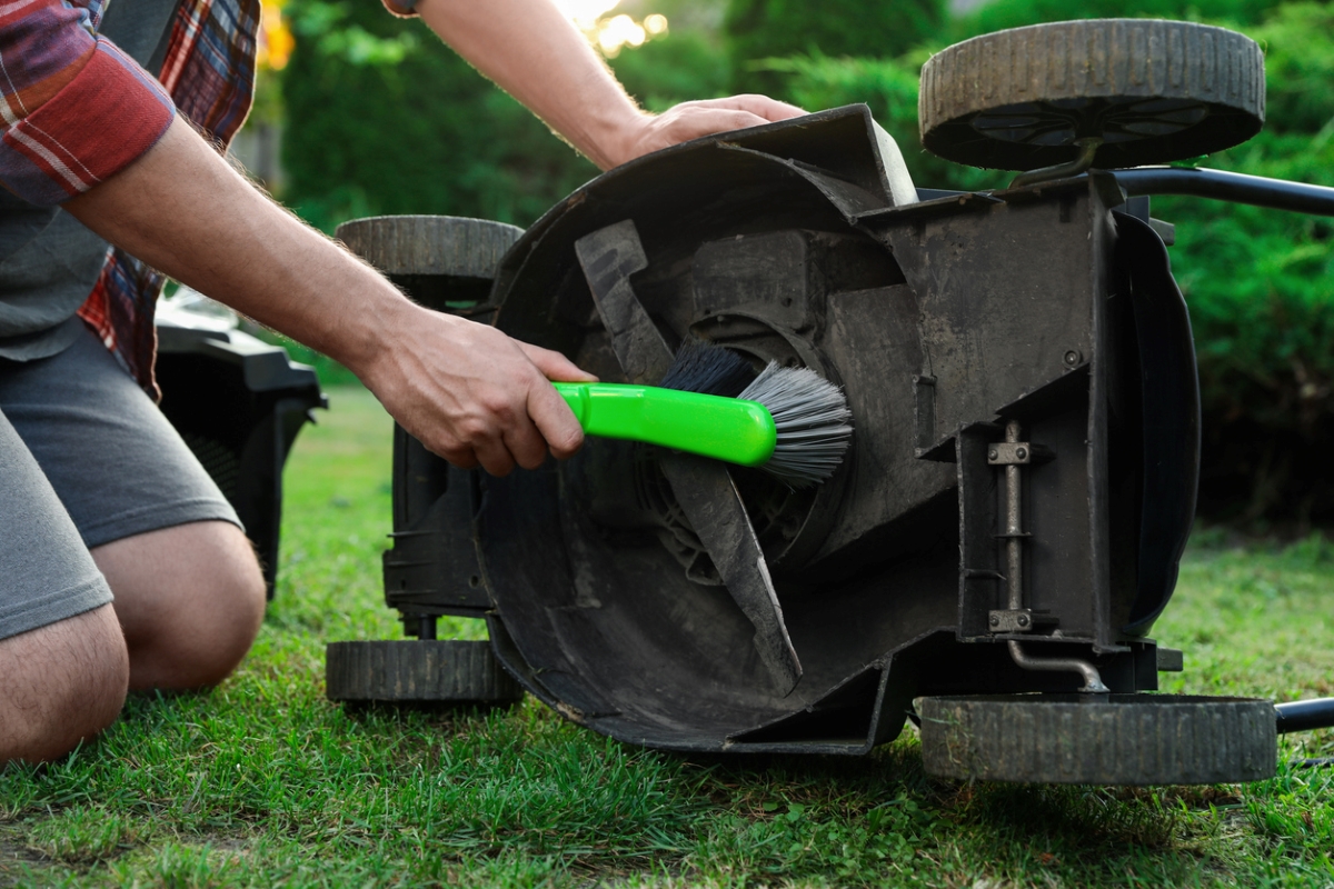 A man kneeling next to flipped lawnmower and brushing the underside.