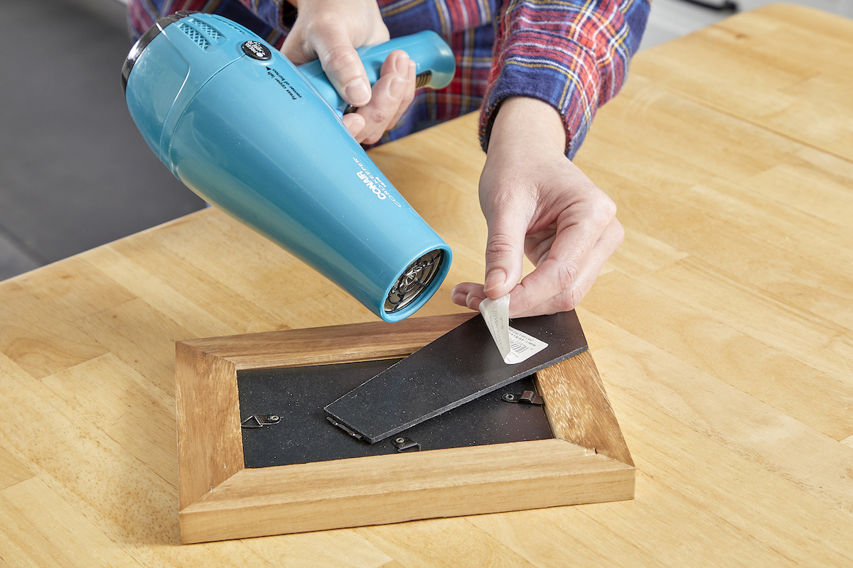 Woman using hair dryer to remove a sticker on a picture frame.