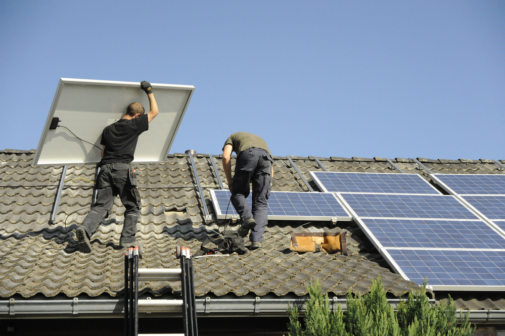 Two workers install solar panels on a tiled roof.