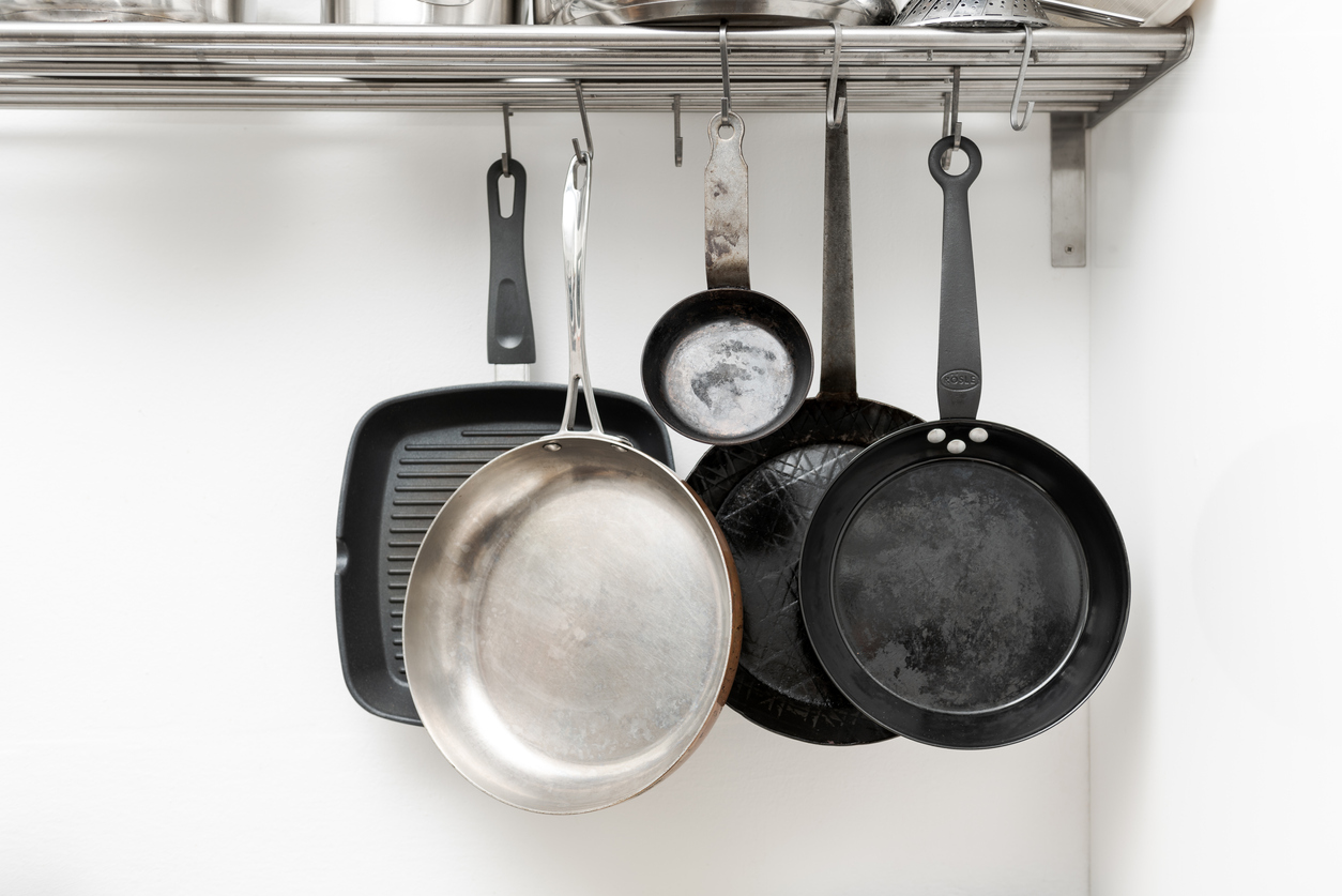 Various pans in different sizes and forms for cooking and frying hanging on metal hooks from shelf in kitchen with white wall in background