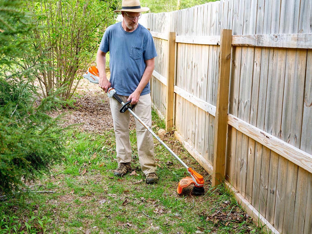 Personne en chemise bleue utilisant un coupe-herbe orange pour couper l'herbe le long d'une clôture en bois