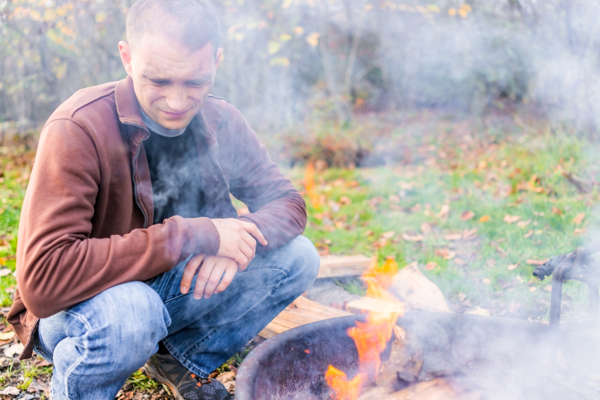 A man is making a face because smoke from a fire pit is blowing into his face.