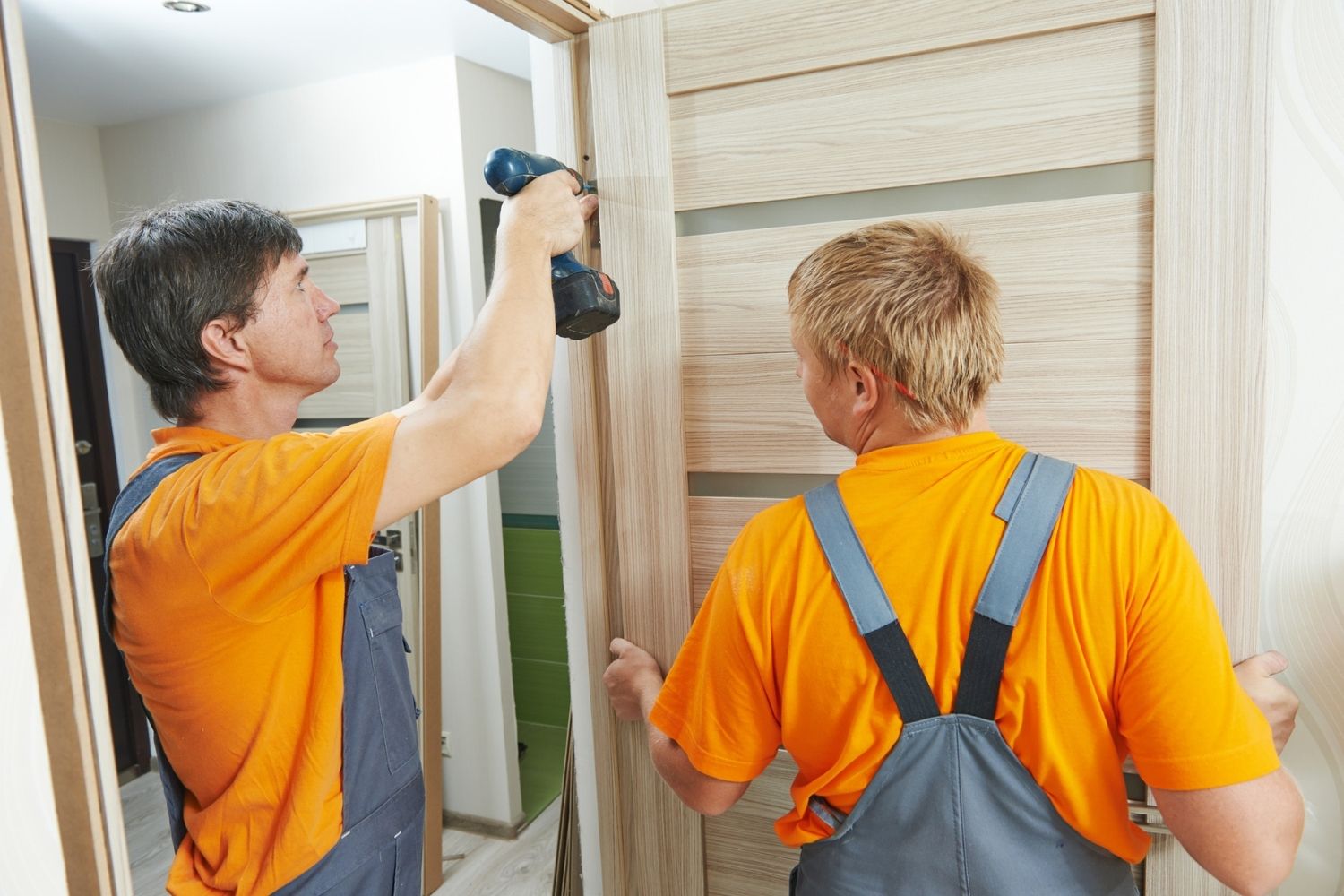 Two workers in yellow shirts and overalls install an exterior door. 