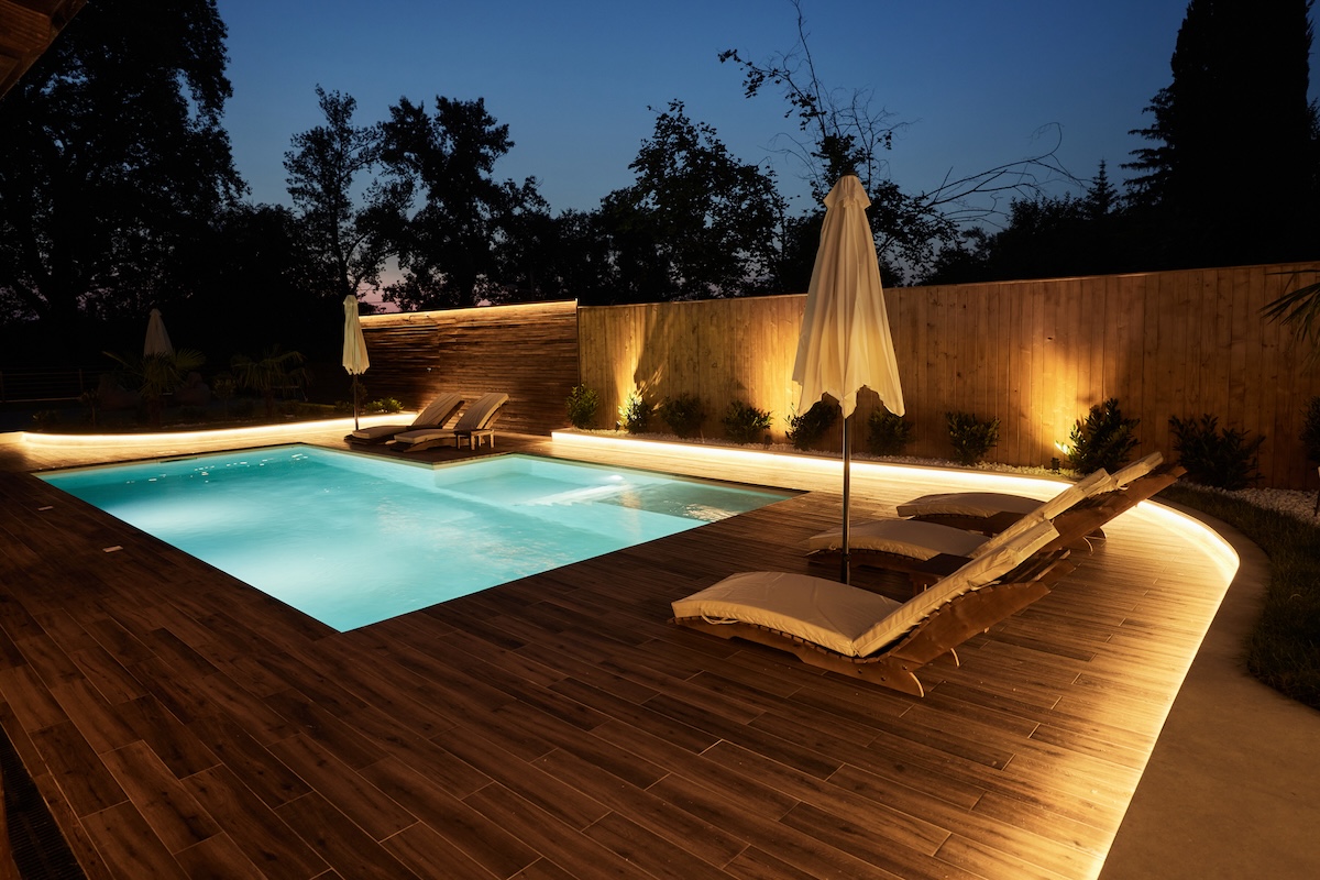 Modern wooden pool deck with brightly-lit swimming pool, deck furniture, and umbrella.