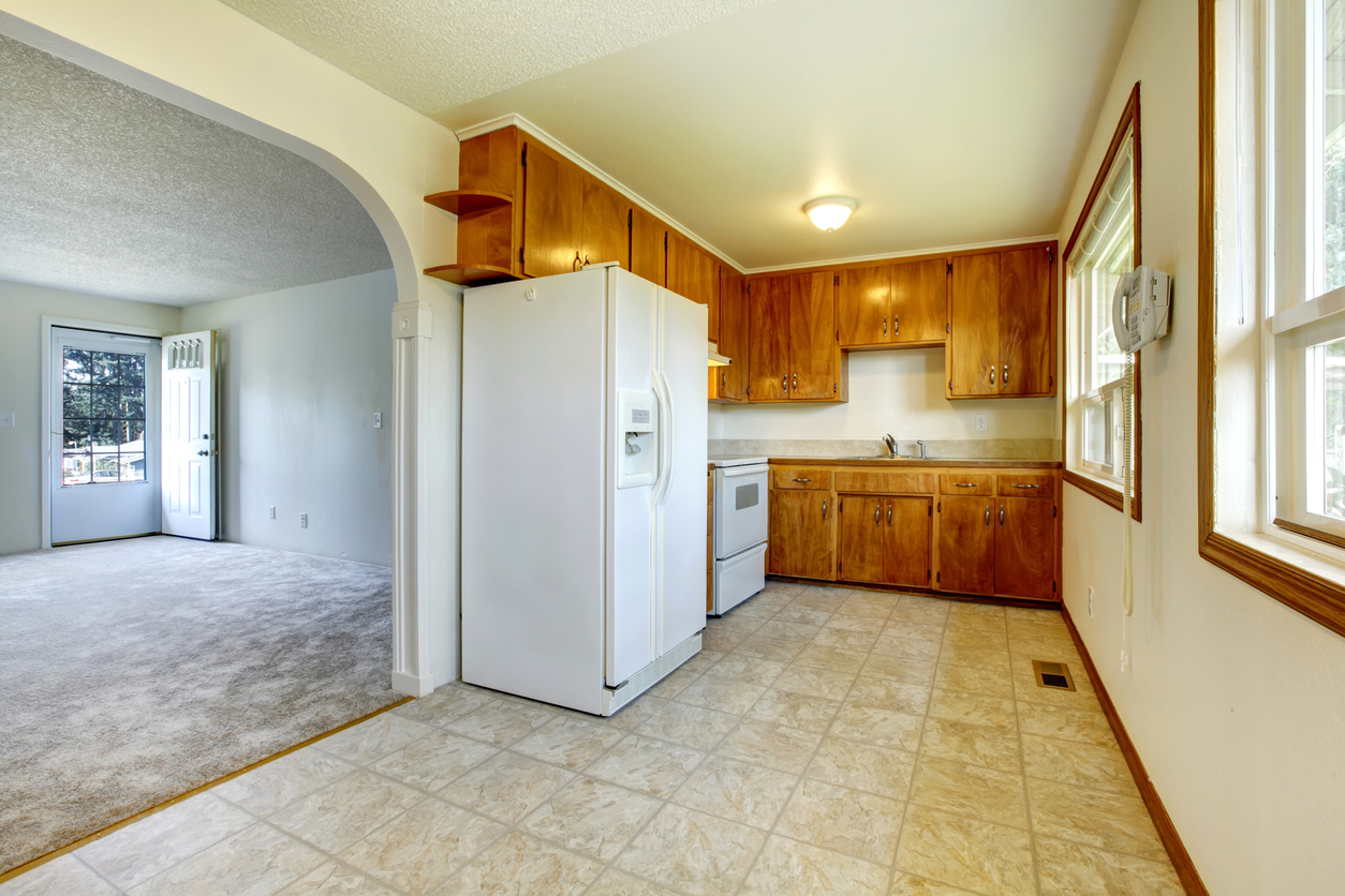 View of the empty living room from a small kitchen with concrete floor, white appliances and wooden cabinets