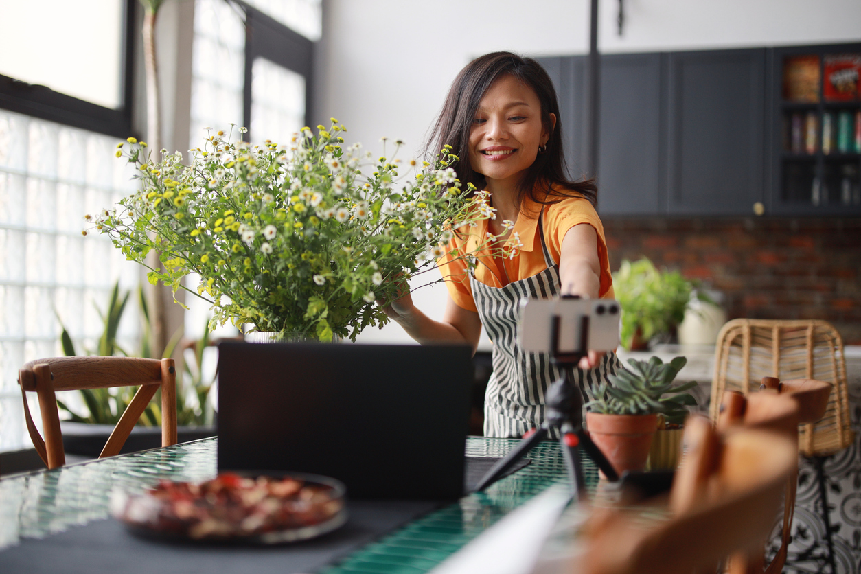 A woman florist is vlogging on her smartphone and laptop, creating an online tutorial on flower bouquet arrangement at home.