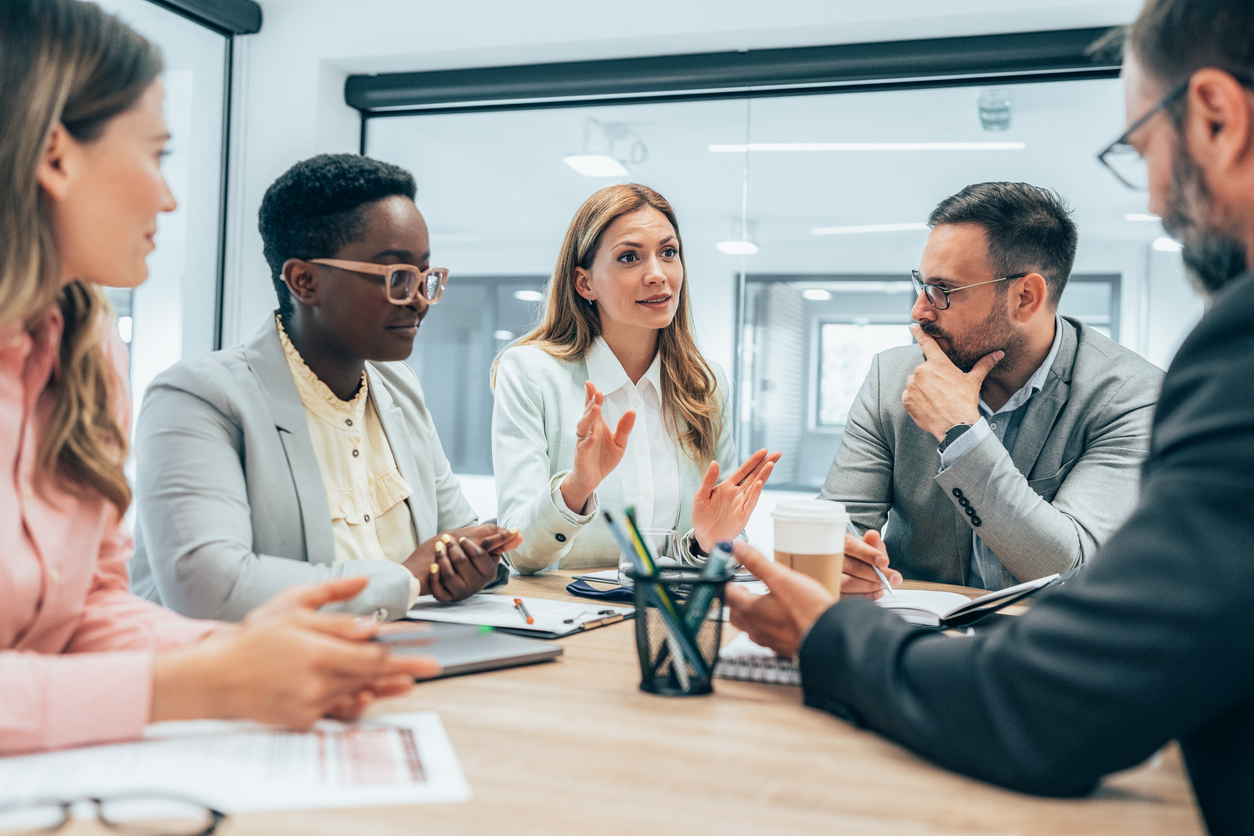 A-group-of-people-sit-at-a-table-and-a-woman-is-speaking-while-the-others-listen.