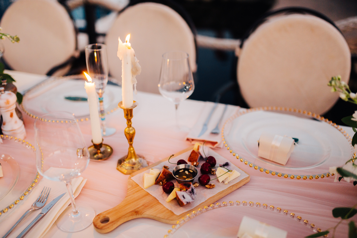 Table decorated with plates with beads and candles on a white tablecloth and cherries and cheese on a cutting board.