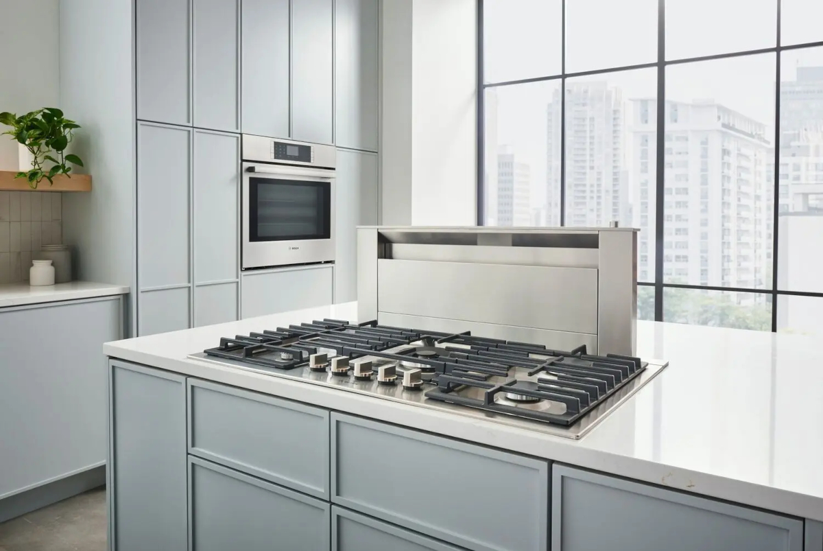 A stainless steel countertop range hood on a kitchen island with a city view outside of the window.