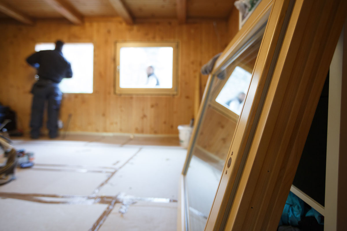 A window is on the ground in the foreground, waiting to be installed by a worked dressed in blue in the background.