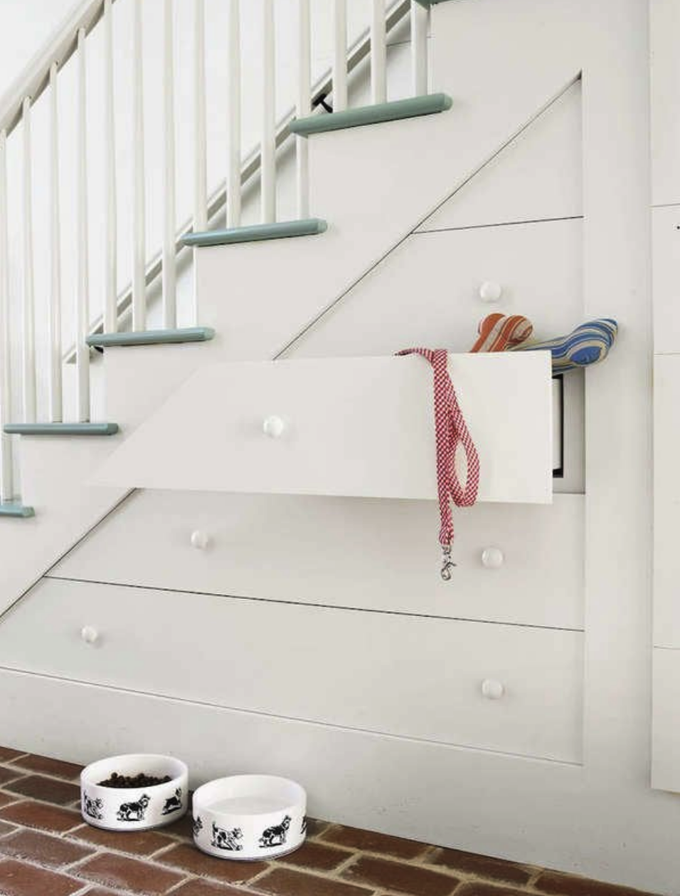 white nook under staircase with green stairs with drawers containing dog leash and toys with dog food and water bowls on the floor