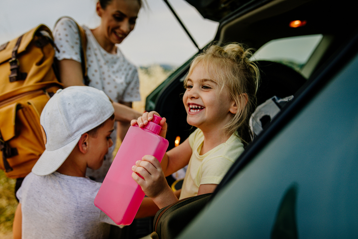 une petite fille est assise dans le coffre d'un hayon et boit dans une bouteille d'eau rose pendant que sa mère et son frère chargent le coffre pour un voyage en voiture