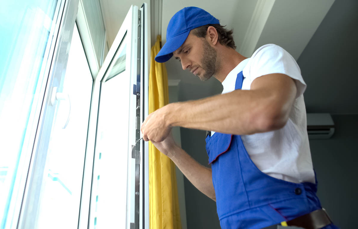 A worker in a blue hat and overalls uses a screwdriver to fix a window frame in a home.