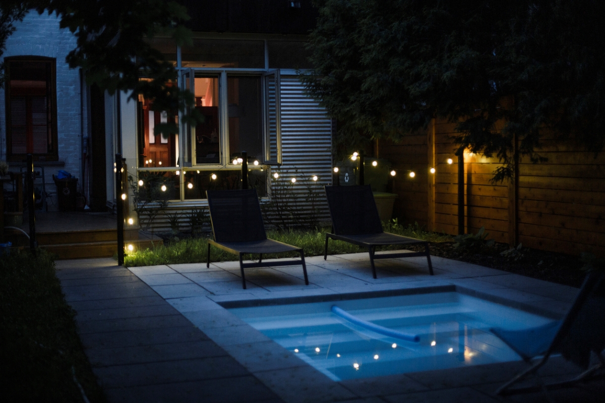 A swimming pool near a home with string lights hanging above. 